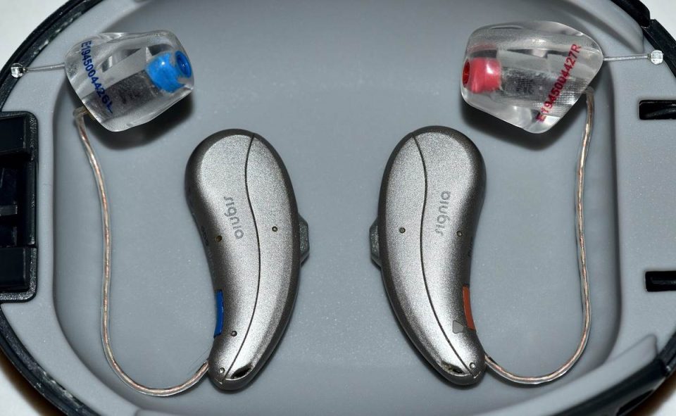 Deux aides auditives en argent avec embouts transparents dans un boîtier gris